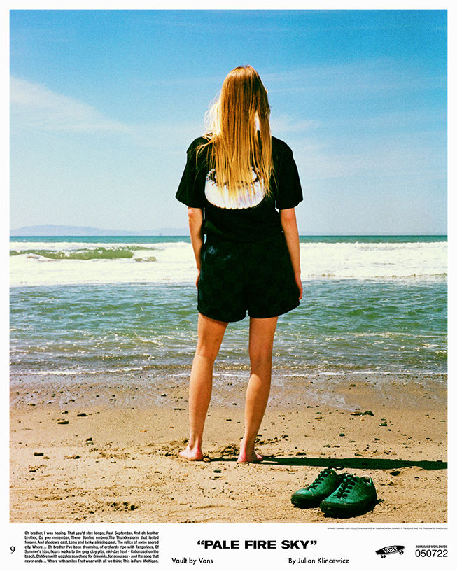 Girl in black shorts and tee shirt, barefoot with back facing camera looking towards the ocean. A pair of teal Vans Authentic shoes are resting on the sand