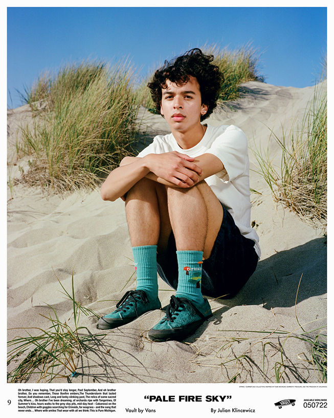 Man sitting on beach sand with white tee shirt, black shorts, and teal Vans Authentic shoes