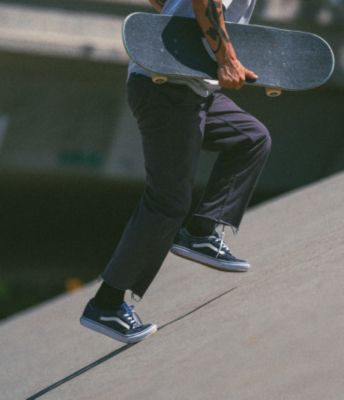 A skateboarder walking uphill while holding his skateboard and wearing Vans Skate Rowley shoes.