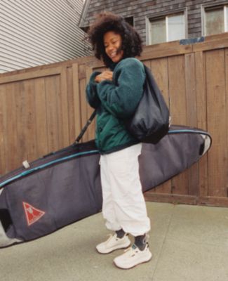 A girl wearing a fleece jacket and white track pants wearing Vans UltraRange holding a surfboard bag.