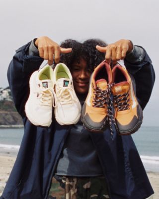 A person holding two pairs of Vans white and orange Crosspath shoes at the beach.