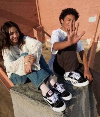 Two big kids smiling together while sitting outside and wearing a pair black and white knu skool shoes and checkerboard knu skool shoes.