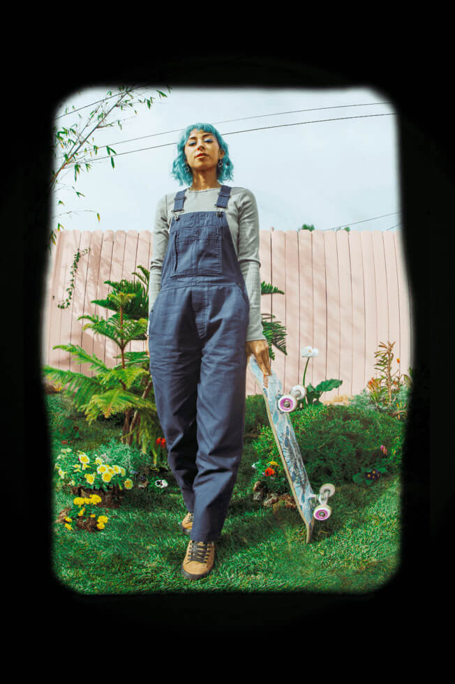 Lizzie Armanto standing with her skateboard and wearing blue overalls from The Lizzie collection.