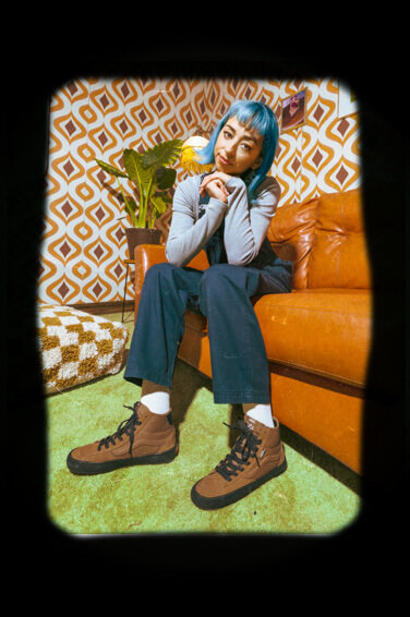 Lizzie Armanto sitting on an orange couch wearing overalls and shoes from The Lizzie collection.